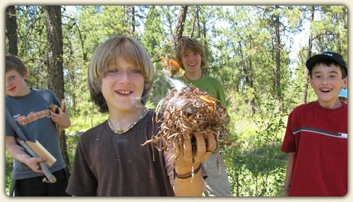 Teenage boy holding dry foliage for fire-starting materials and flames coming from the top, as another boy looks on from behind him. Fire and shelter building, ethics/Leave No Trace, and biking skills are just a few of the learning adventures that await your child. Photo: Twin Eagles Wilderness School