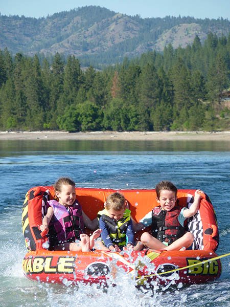 Three kids sitting on a large orange inflatable tube-seat, being pulled by a powerboat, and kids smiling with excitment as they are pulled fast with lake water spraying in their faces.