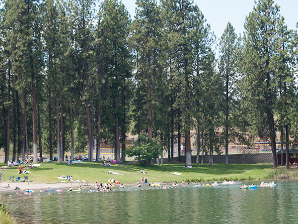 Medical Lake's Waterfront Park with a sandy beach. People swimming and lounging on the grass, with tall Ponderosa pine trees in the background.