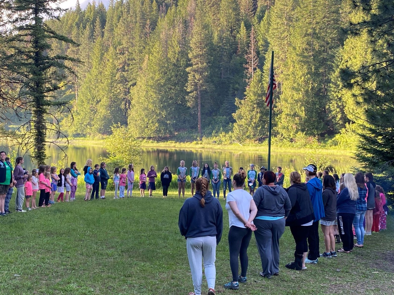 Girl Scout summer campers standing in a big circle on the grass, with Lake Coeur d'Alene and trees in the background.