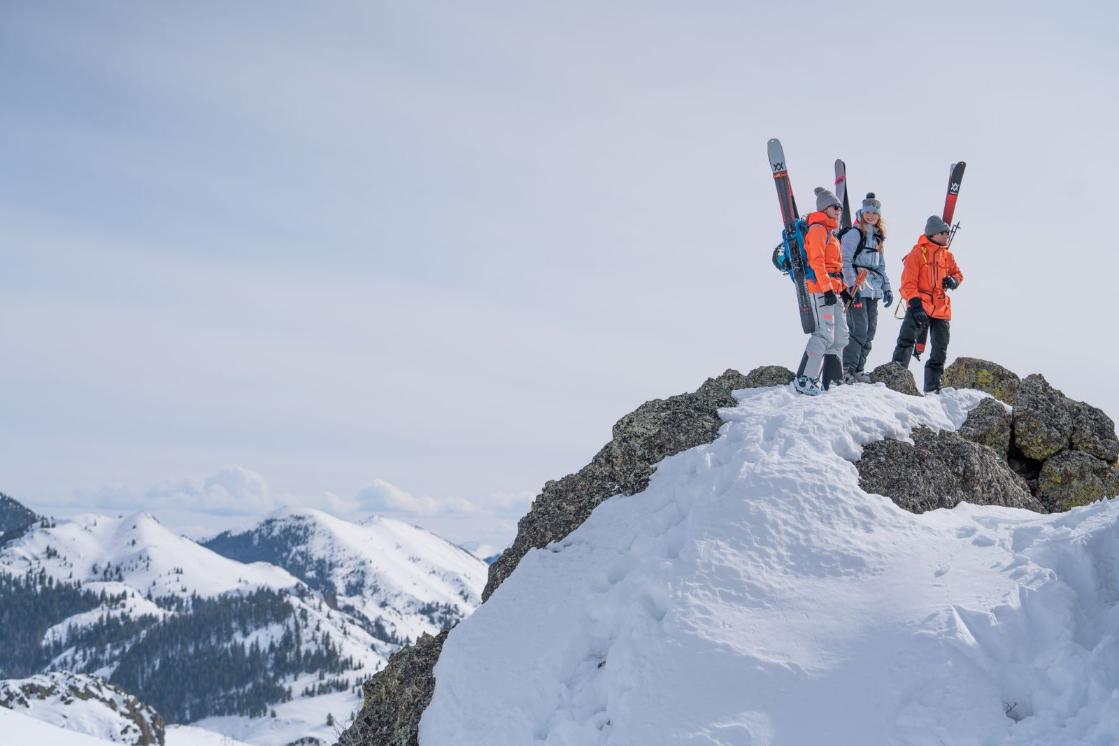 Three skiers in their ski boots with skis attached to their backpacks as they stand atop a rock cliff on a mountain.