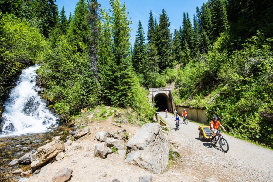 People biking on trail, outside an old train tunnel, with a small waterfall off to the side.