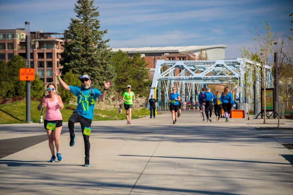 A group of runners on a race course through Riverfront Park in downtown Spokane, after they've passed over a steel structure bridge over the Spokane River. A male runner gleefully smiles and waves to the camera while hopping in the air.