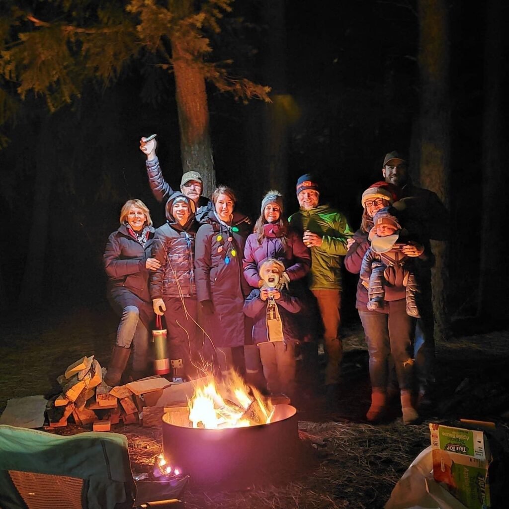 Group of people wearing winter coats, hats, and gloves standing around a campfire and smiling at the camera.