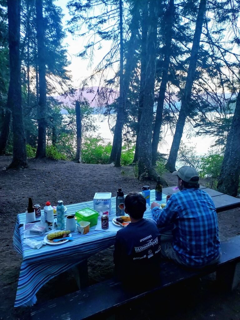 View of campsite picnic table and family eating dinner, near dusk, with darkened view pine trees and bushes close by, with view of lake below.