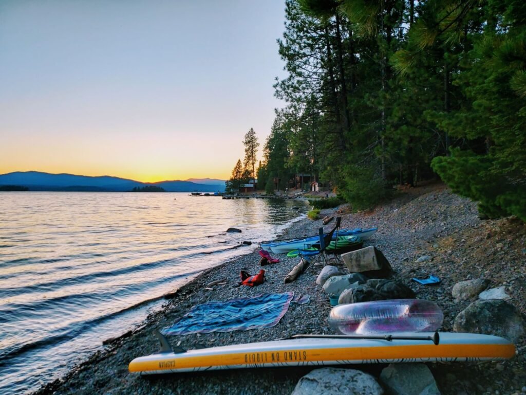 Beach base camp with stand-up paddleboard, two kayaks, life jackets, blanket, tube, and chair at Sam Owen Campground along Lake Pend Oreille, with orange and yellow sunset on the horizon in the far distance. // Photo: Amy McCaffree