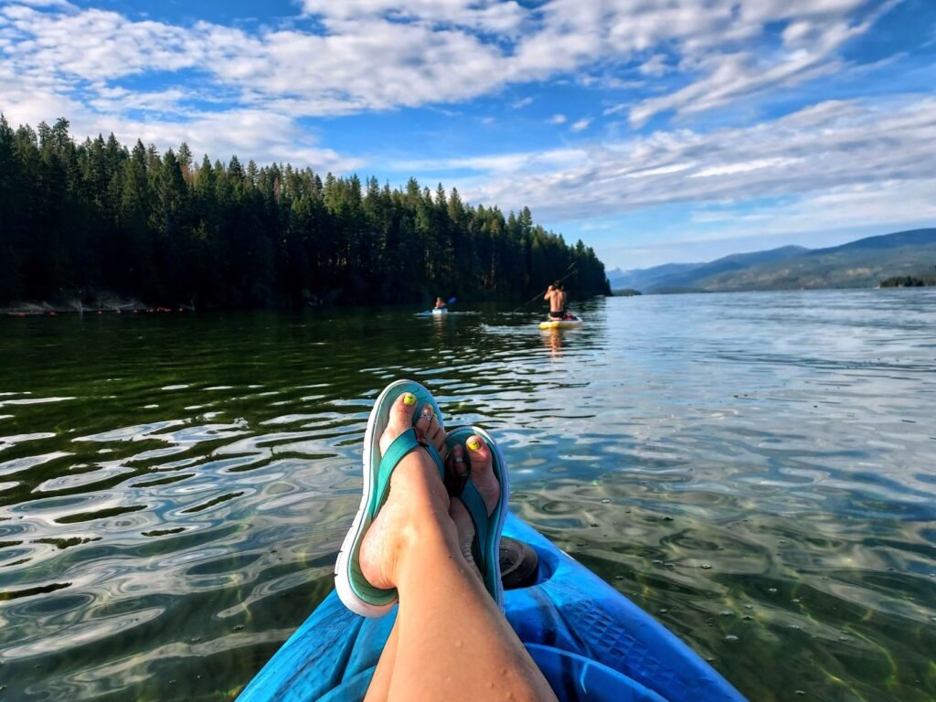 Kayaker's perspective view of paddling on Priest Lake, with her sandaled feet propped up kayak bow with view of children paddling nearby, one in a kayak and one on a stand-up paddleboard