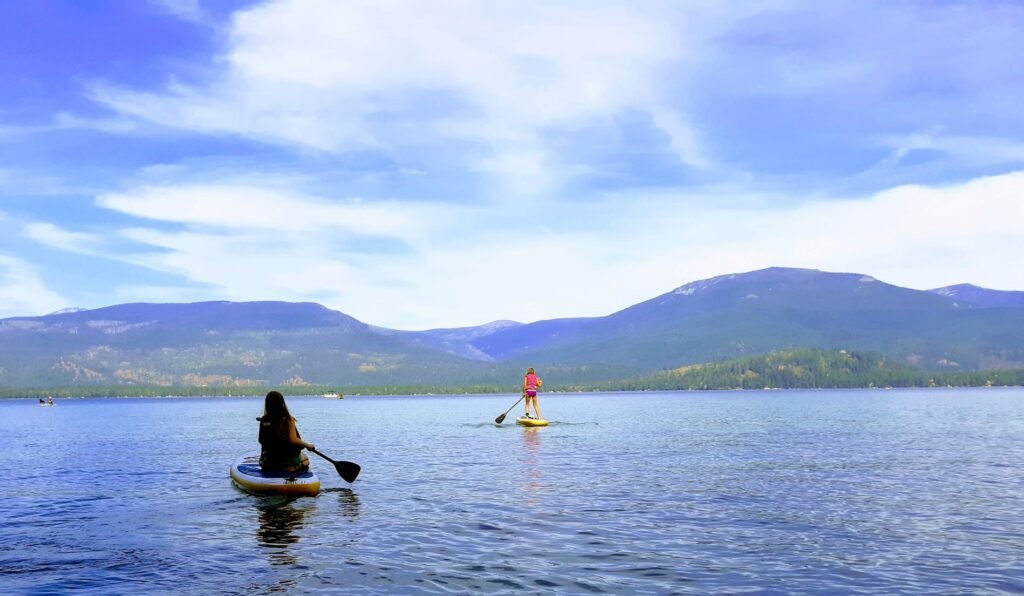 Blue-colored lake water and blue sky with streaks of white clouds and two people on stand-up paddleboards, paddling on on Priest Lake, with view of hills in the far distance on the other side of lake.