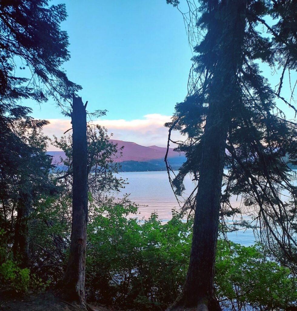 View of Priest Lake beyond campsite pine trees, near sunset time, with pink and purple hues on water and on the hills on the opposite shore. 