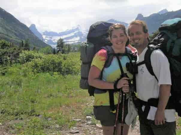 Amy and her husband wearing their backpacks on a trail in Glacier National Park with a view of the park's iconic glacier-carved mountain peaks in the distance.s, after crossing the river using the Cosley Lake Outlet Safety Cable.