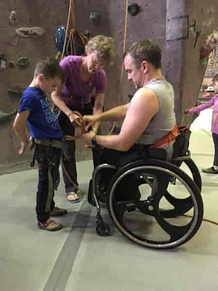 Photo of Tyler Byers in wheelchair, tying a figure-eight through his son's harness.