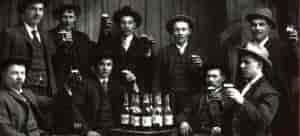 Black and white vintage photo of men in black suits standing next to bottles of beer.