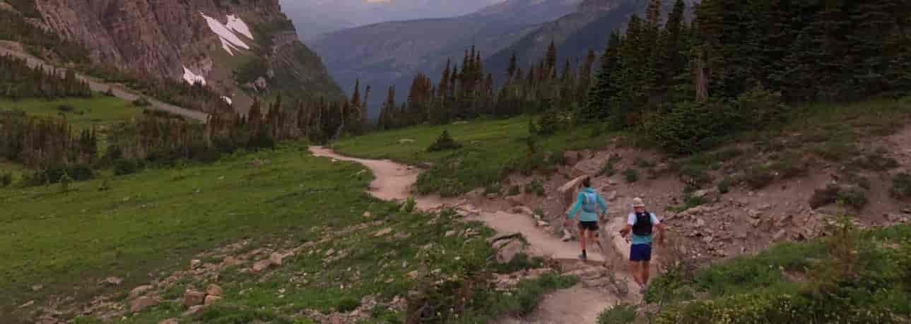 Runner along a singletrack dirt trail in the backcountry, through a meadow, with mountain peaks in the distance.