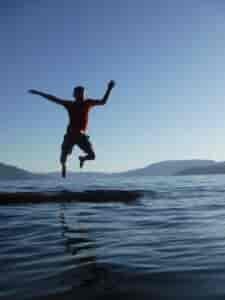 A boy jumping off a log floating in Lake Pend Oreille, with a view of hills in the far background.
