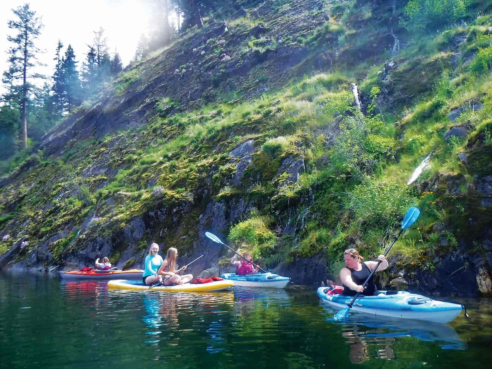 Mom and children paddling in kayaks and a stand-up paddleboard along a rocky slope of Lake Sullivan. Mossy grass and waterfall on the rocks.
