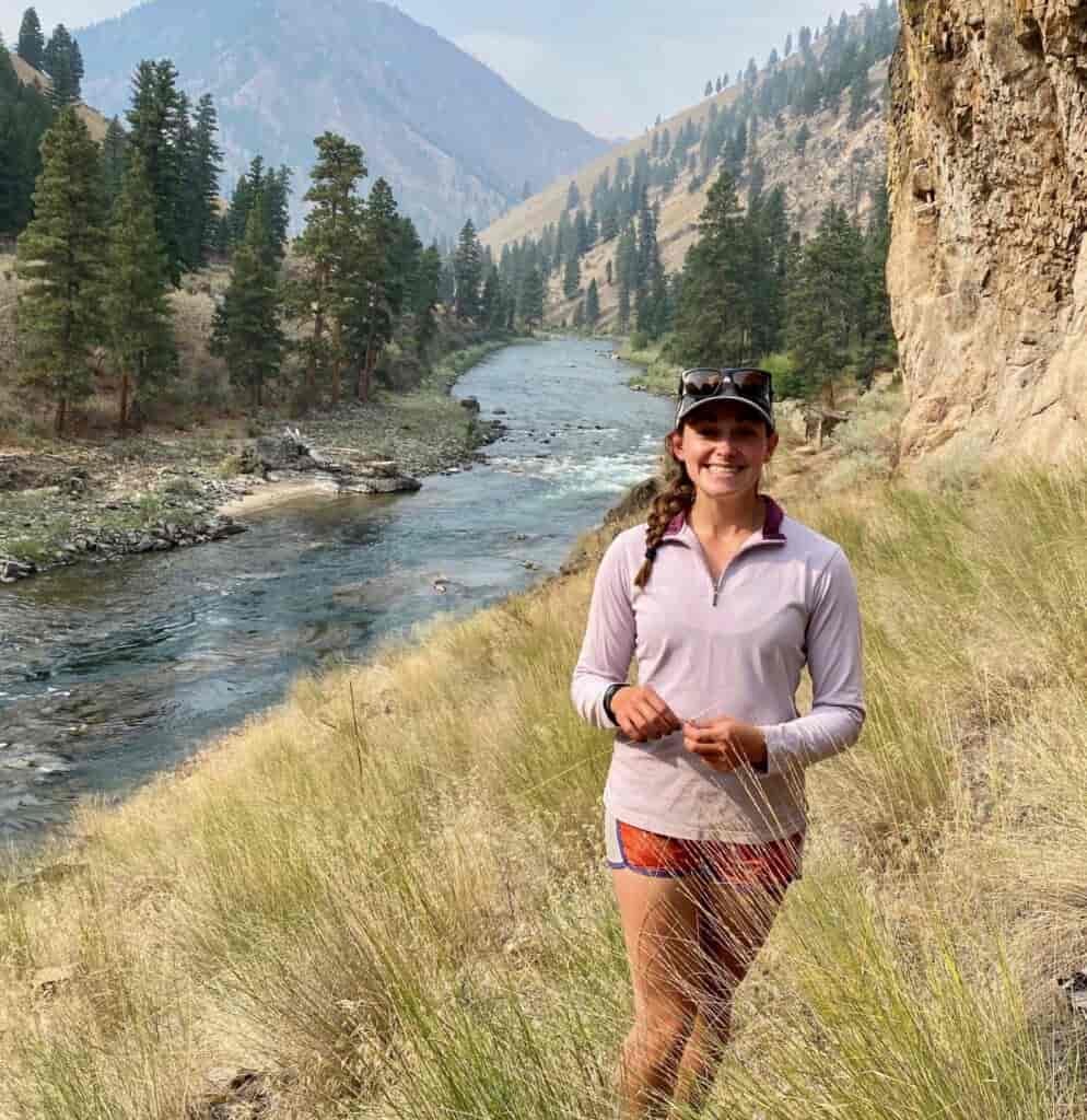 Woman smiling at the camera, standing in a grassy riverbank, with view of river, trees, and hillsides in the background.