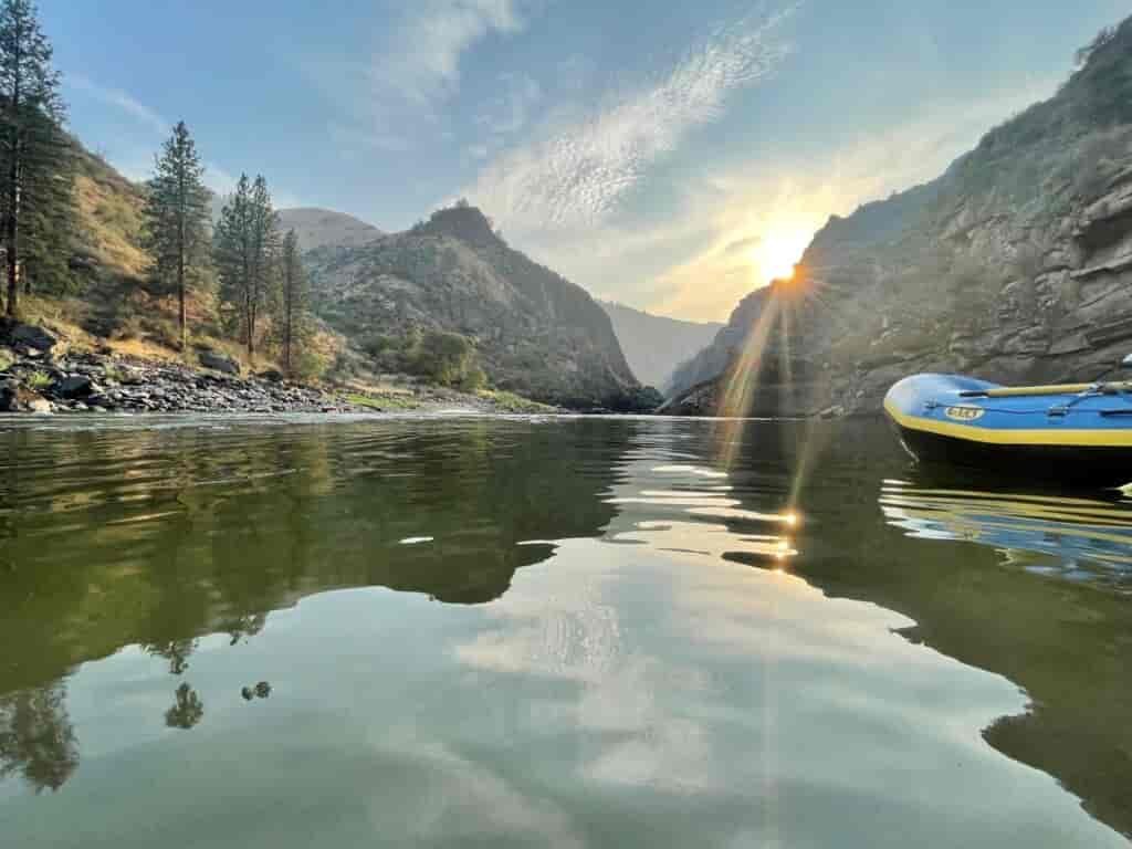Flat water section of the Snake River, with a raft floating on it, and rocky canyon walls with sun party obscured behind them.