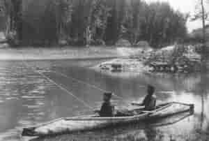 Sepia-tone photo of traditional Kalispel Tribe canoe and two people sitting inside with fishing rods in the water.