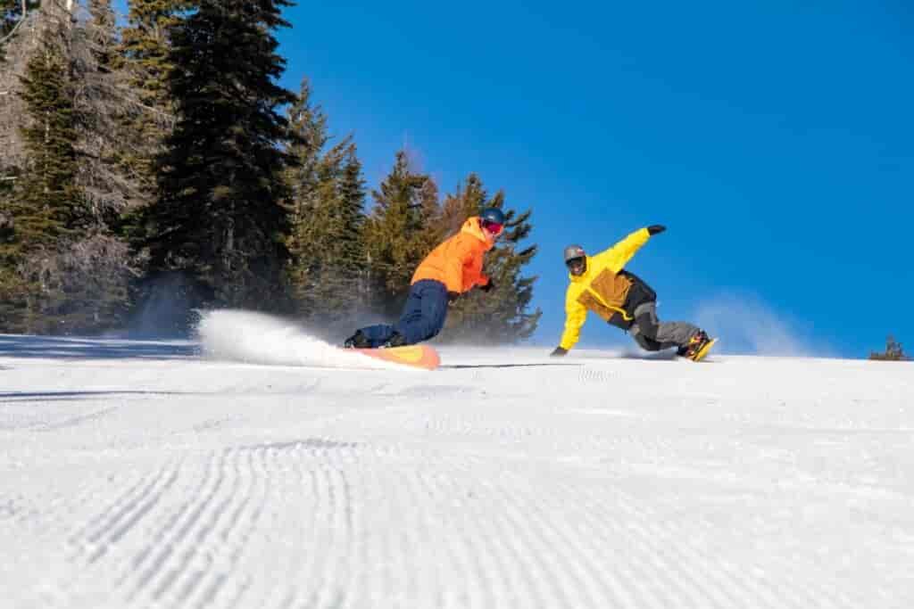Two snowboarders, one wearing an orange jacket and the other in yellow, making big turns down a groomed run at 49 Degrees North.