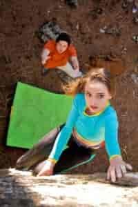 A woman wearing a bright blue long-sleeved shirt and black climbing pants is reaching up for a hand hold on a boulder. Below her lays a green safety mat on the ground and a male climber with arms raised spotting her in case she falls.